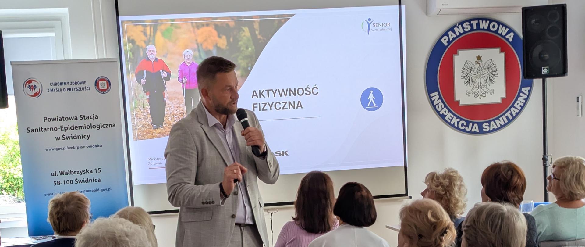 Prelegent prowadzi wykład dla grupy seniorów podczas spotkania edukacyjnego w Powiatowej Stacji Sanitarno-Epidemiologicznej w Świdnicy. Na ekranie wyświetlana jest prezentacja pt. „Aktywność fizyczna”, a uczestnicy siedzą w rzędach i słuchają wystąpienia. W sali widoczne są materiały informacyjne oraz znak Państwowej Inspekcji Sanitarnej.