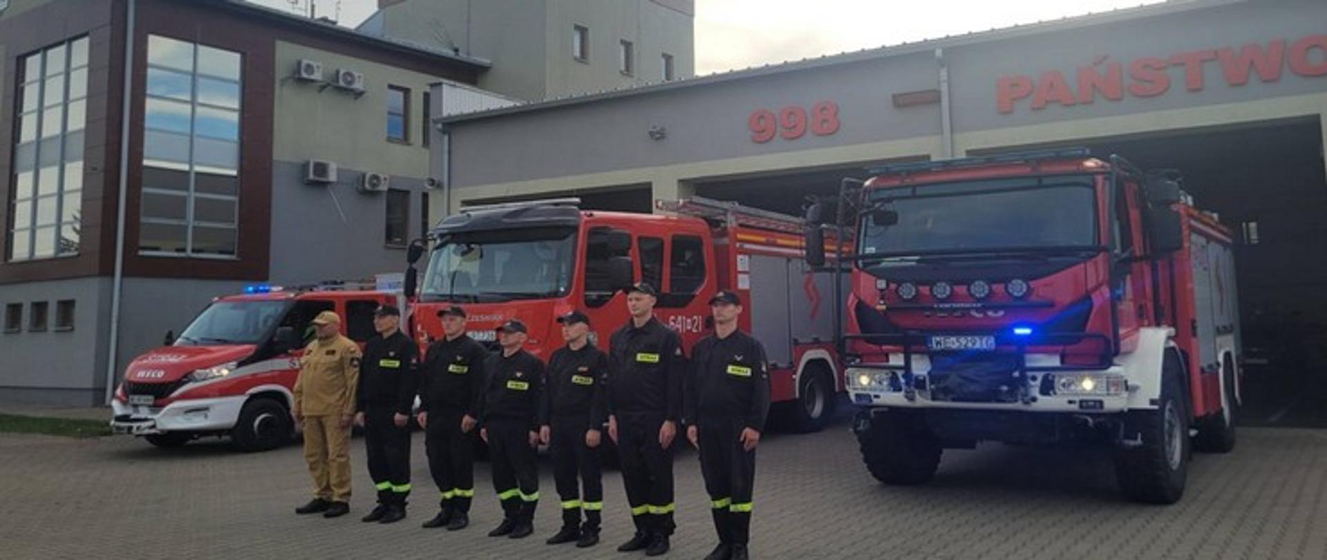 Na zdjęciu widzimy funkcjonariuszy Jednostki Ratowniczo-Gaśniczej w Sokołowie Podlaskim, którzy stoją na baczność. Ubrani w ubrania koszarowe koloru czarnego. Stoją na kostce brukowej przed pojazdami ratowniczymi koloru czerwonego. Dalej widoczny jest budynek Komendy Powiatowej Państwowej Straży Pożarnej koloru szaro-żółtego.
