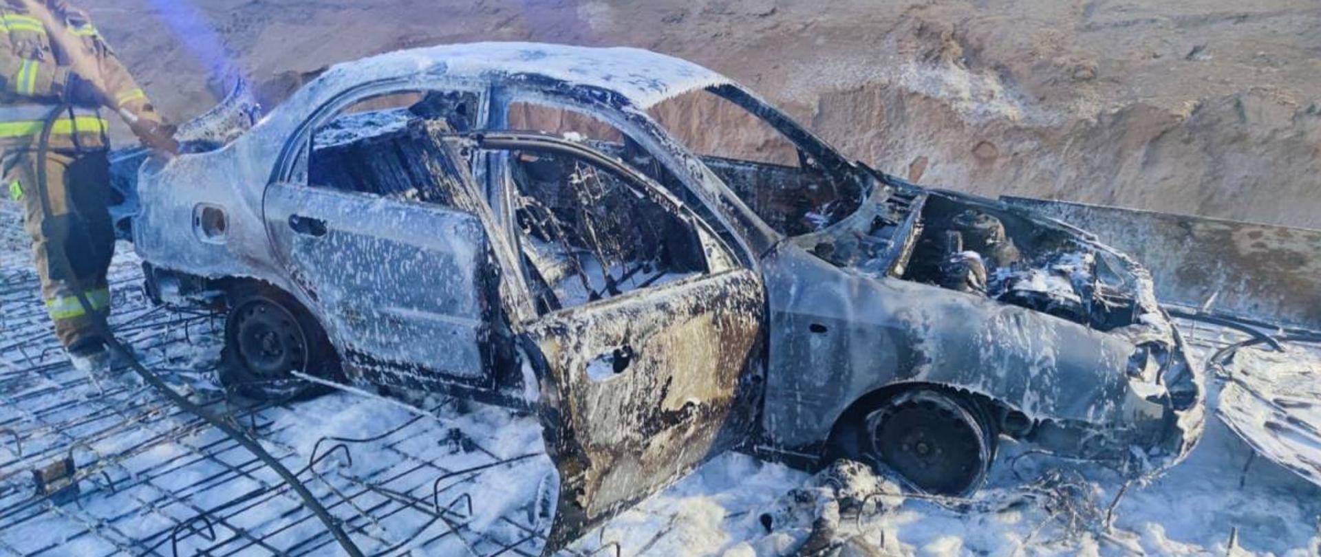 Pokryty pianą gaśniczą spalony wrak samochodu osobowego, strażak z prądownicą.