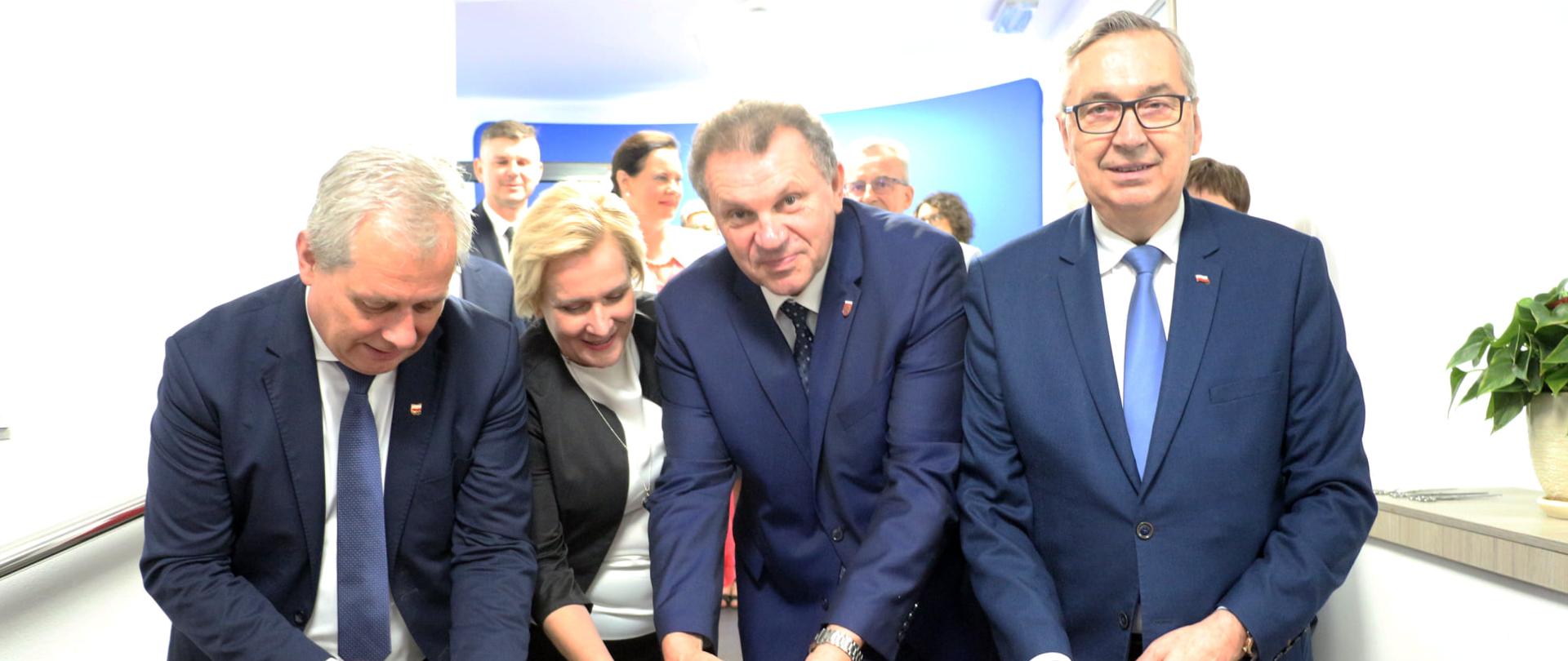 Na zdjęciu cztery osoby przecinające czerwoną wstęgę. W środku kobieta - wicewojewoda opolski. Wszyscy pochylają się nad wstęgą, spoglądają w obiektyw na przeciw - pozują do zdjęcia. W swoich dłoniach mają nożyczki, przecinające wstęgę. W tle ścianka z napisem GOGOLIN.