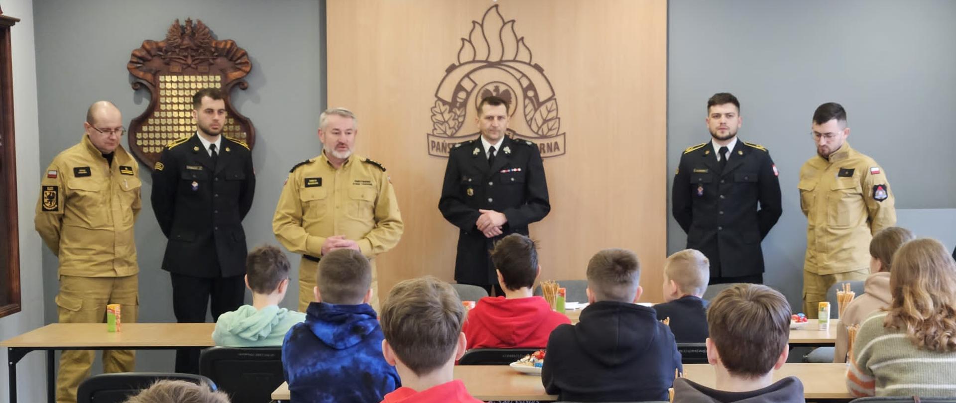 Świetlica komendy. Dzieci siedzą w ławkach w tyle strażacy PSP i OSP. Komendant wita dzieci