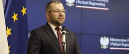 Minister funduszy i polityki regionalnej Grzegorz Puda przemawia do mikrofonu, za jego plecami flagi Polski i UE oraz baner MFiPR