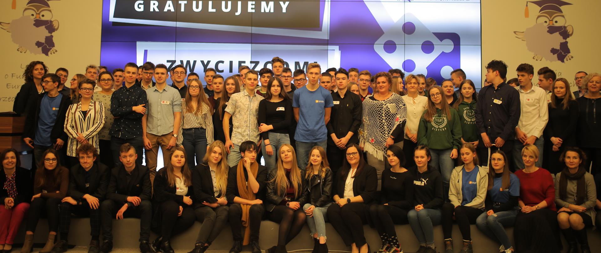 zdjęcie grupowe wszystkich finalistów, którzy stoją lub siedzą na stopniu sceny i patrzą w stronę fotografującego