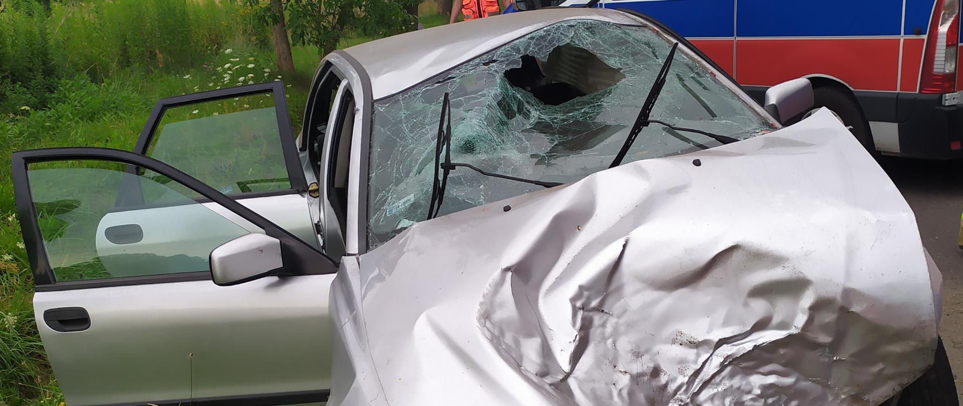 Na środku zdjęcia widać rozbity samochód osobowy marki volvo koloru srebrnego który stoi na poboczu drogi. W pojeździe uszkodzone zostały przednia maska, która jest pognieciona i zdeformowana, rozbita przednia szyba od strony pasażera oraz kierowcy. Uszkodzeniu uległ również dach pojazdu , który jest pogięty. W pojeździe brakuje przedniego zderzaka oraz lamp, które w wyniku zdarzeniu odpadły z pojazdu. Przednie drzwi od strony pasażera i tylnie za pasażerem są otwarte.
Przed pojazdem na poboczu znajduje się zielona trawa. Po prawej stronie zdjęcia za pojazdem stoi karetka pogotowia ratunkowego koloru biało niebiesko czerwonego z włączonymi sygnałami świetlnymi. Przy przedniej części karetki widać ratownika medycznego w maseczce ochronnej na twarzy i czerwonym stroju. W oddali widoczne są rosnące liściaste drzewa.