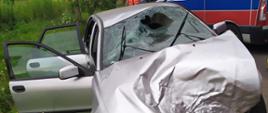 Na środku zdjęcia widać rozbity samochód osobowy marki volvo koloru srebrnego który stoi na poboczu drogi. W pojeździe uszkodzone zostały przednia maska, która jest pognieciona i zdeformowana, rozbita przednia szyba od strony pasażera oraz kierowcy. Uszkodzeniu uległ również dach pojazdu , który jest pogięty. W pojeździe brakuje przedniego zderzaka oraz lamp, które w wyniku zdarzeniu odpadły z pojazdu. Przednie drzwi od strony pasażera i tylnie za pasażerem są otwarte.
Przed pojazdem na poboczu znajduje się zielona trawa. Po prawej stronie zdjęcia za pojazdem stoi karetka pogotowia ratunkowego koloru biało niebiesko czerwonego z włączonymi sygnałami świetlnymi. Przy przedniej części karetki widać ratownika medycznego w maseczce ochronnej na twarzy i czerwonym stroju. W oddali widoczne są rosnące liściaste drzewa.