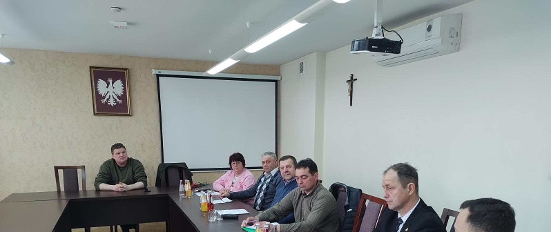 Grupa ludzi, jedna kobieta i siedmiu mężczyzn siedzą przy stole. Na stołach znajdują się butelki z wodą oraz szklanki a także papiery i dokumenty. Znajdują się w jasnym pomieszczeniu, ściany mają kolor jasnego beżu. Z sufitu zwisa rzutnik.