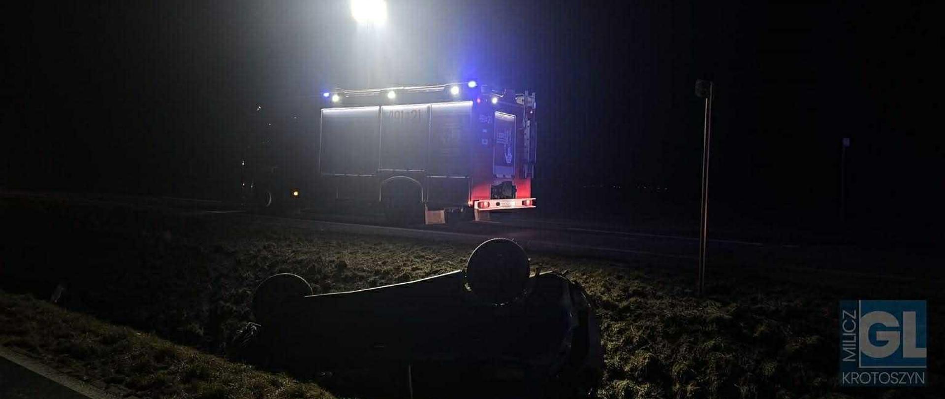 Zdjęcie przedstawia samochód osobowy w przydrożnym rowie oraz wóz strażacki z włączonymi niebieskimi światłami błyskowymi.