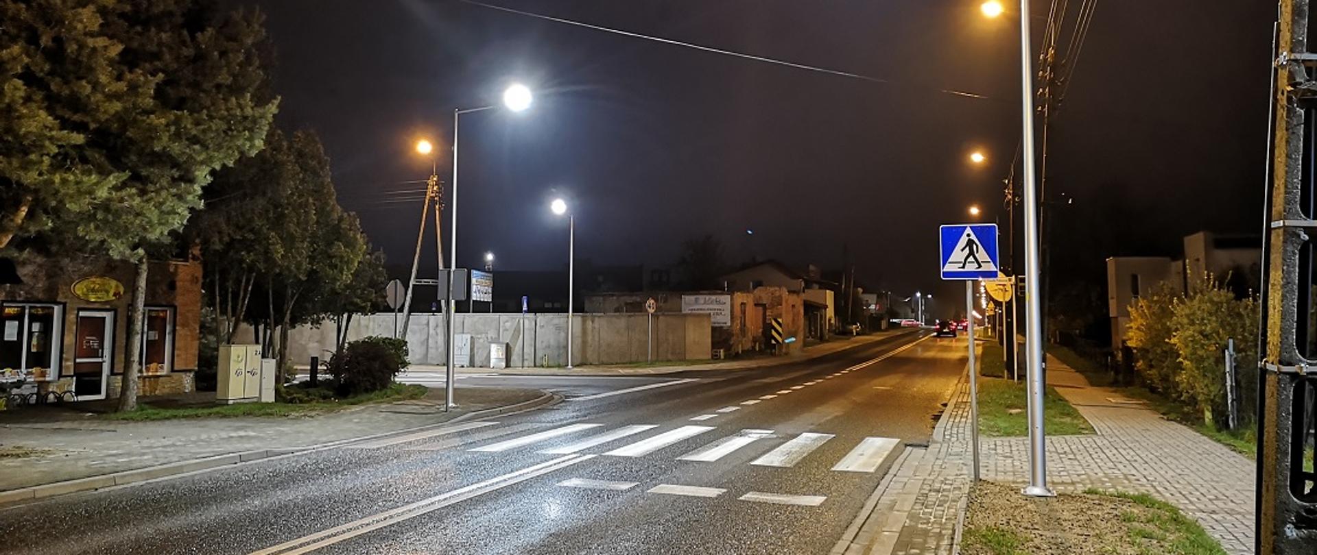 Przejście dla pieszych w terenie zabudowanym nocą. Widoczne oświetlenie uliczne oraz lampy doświetlające samo przejście.