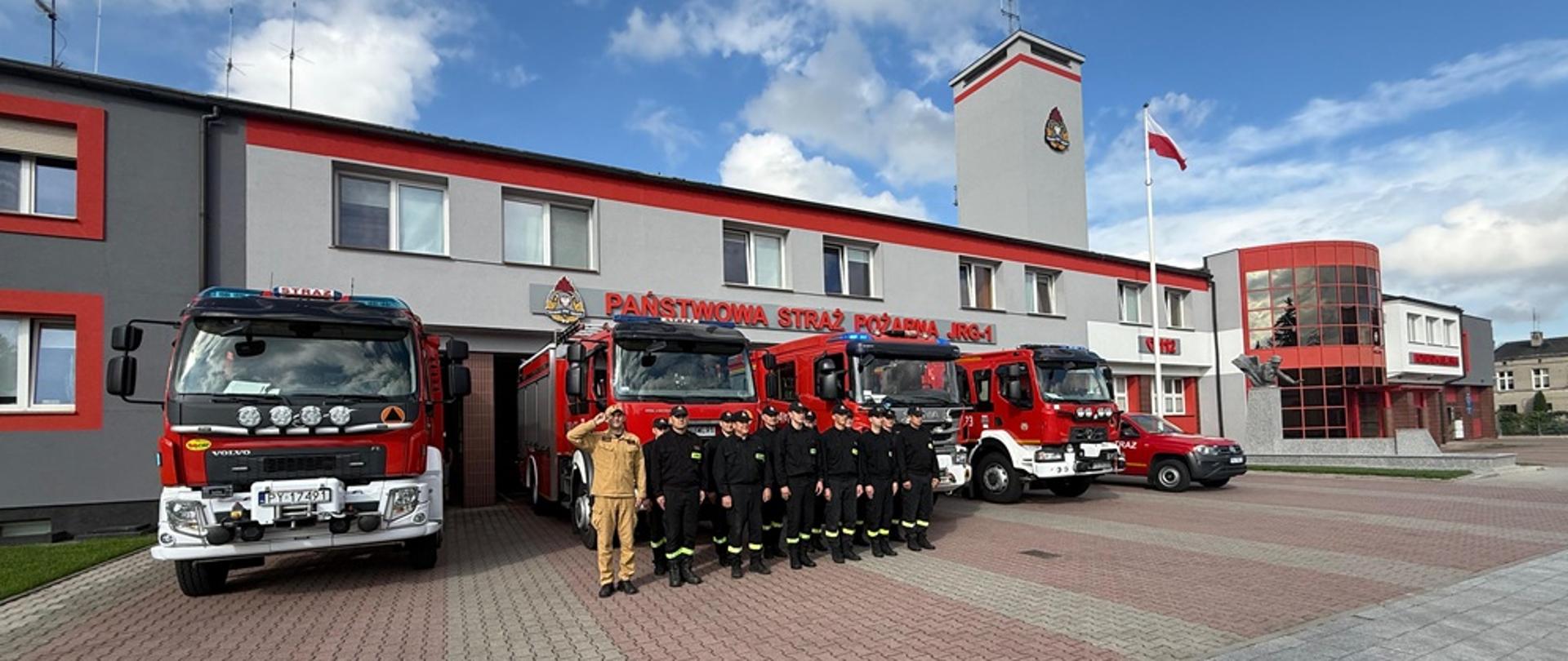 Strażacy w dwuszeregu na tle samochodów strażackich.