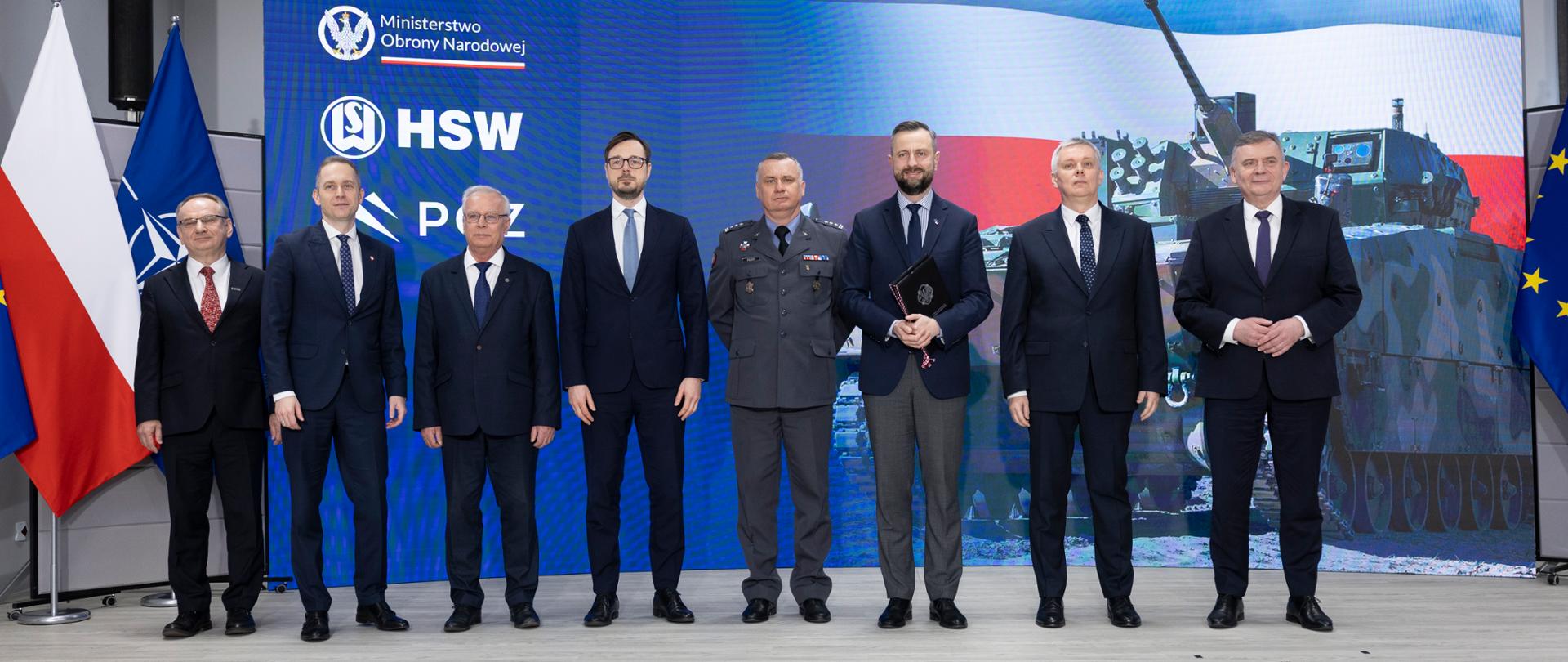 Grupowe zdjęcie ośmiu mężczyzn w garniturach i mundurze wojskowym, stojących na tle niebieskiej planszy z logotypami MON, HSW i PGZ oraz grafiką przedstawiającą sprzęt wojskowy. Po lewej stronie widoczne flagi Polski i NATO.