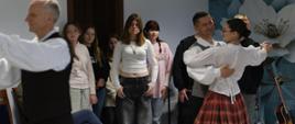 Tańcząca para w stroju ludowym, przy ścianie stoi grupa przyglądającym się dziewcząt.