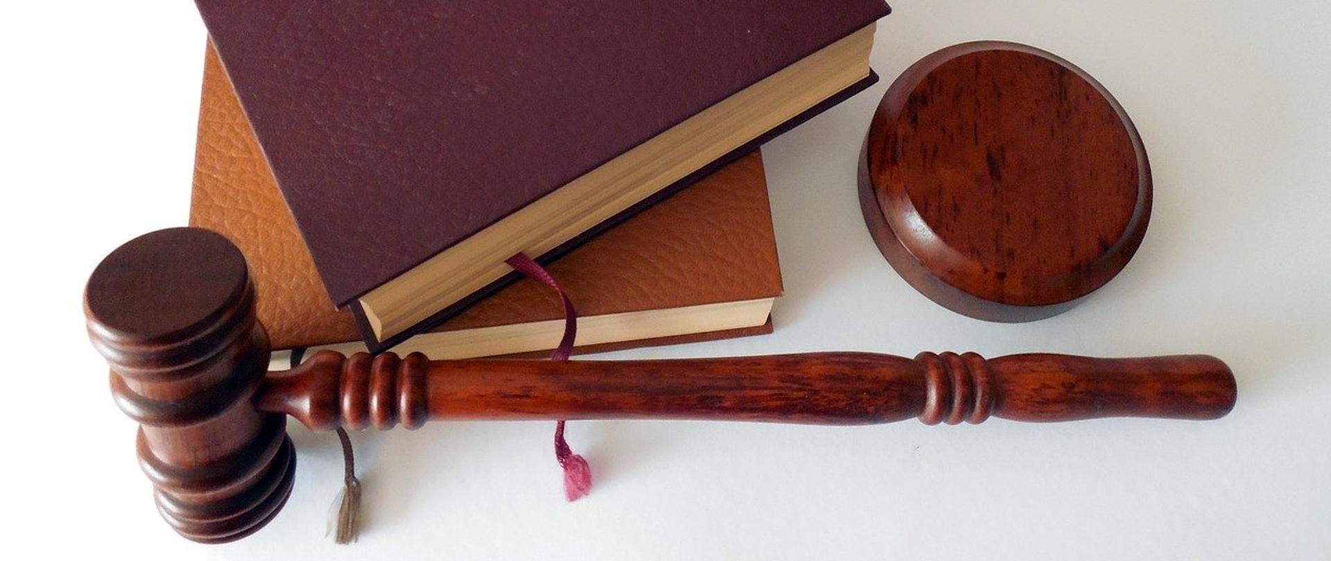 Widok z góry na biały stół. Na stole leżą dwie grube księgi w skórzanych, brązowych oprawach z wystającymi materiałowymi zakładkami. Obok nich znajduje się młotek sędziowski z podstawką.