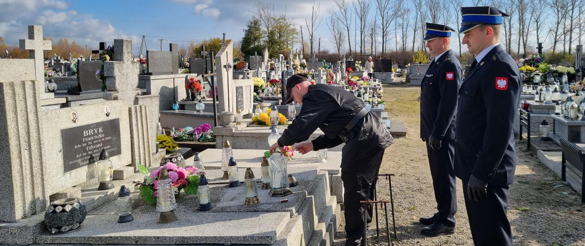 Zdjęcie zrobione na cmentarzu. Przedstawia trzech strażaków nad grobem zmarłego kolegi. Jeden z funkcjonariuszy zapala symboliczny znicz nad mogiłą.