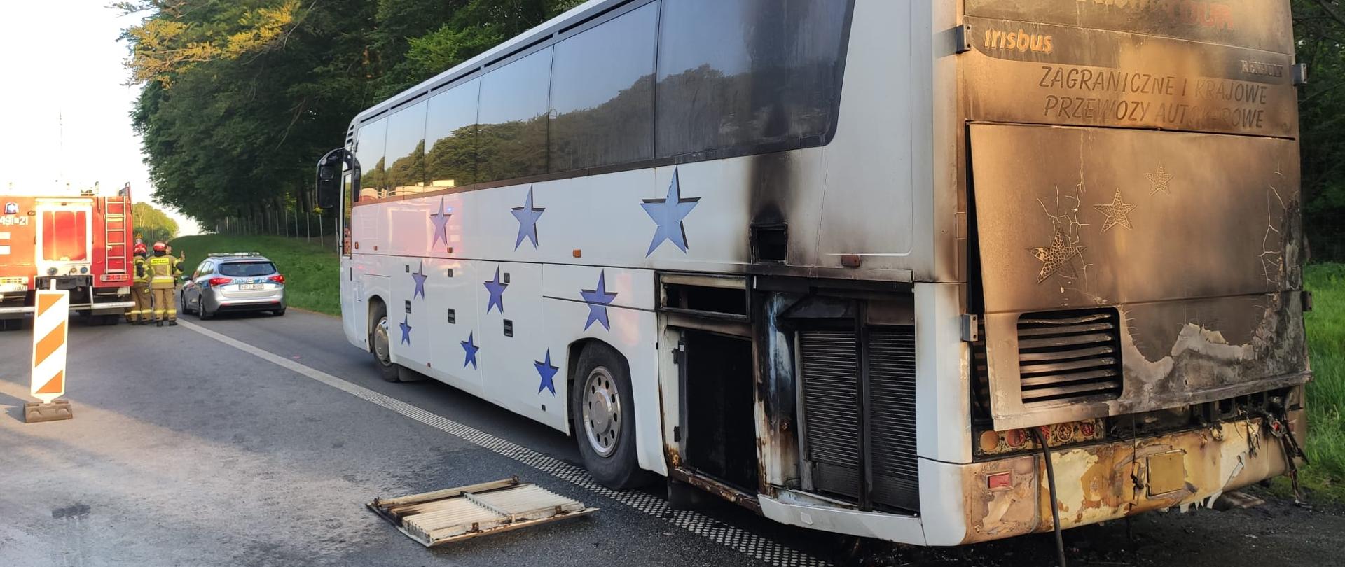 Zdjęcie przedstawia autokar z okopconą karoserią stojący na autostradzie na tle drzew, w głębi widać strażaków i policjantów przy swoich pojazdach