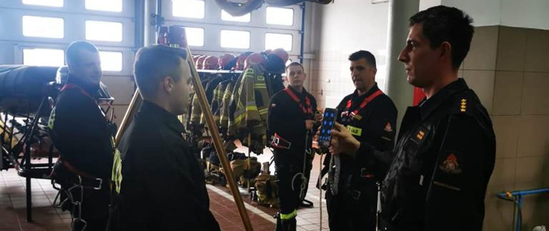 Garaż Jednostki Ratowniczo-Gaśniczej, strażacy uczestniczący w zajęciach doskonalenia zawodowego z ratownictwa wysokościowego. Wykorzystują trójnóg oraz sprzęt wysokościowy i sprzęt ochrony osobistej 