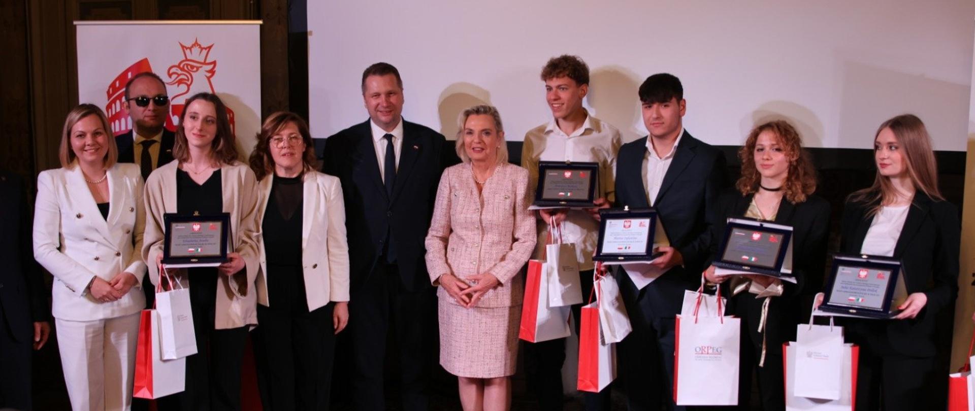 Pod jasną ścianą stoi minister Czarnek w otoczeniu kilku osób, kilkoro z nich trzyma oprawione w ramki dokumenty i biało-czerwone papierowe torby.