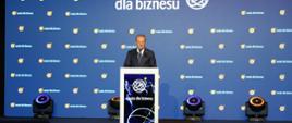 Na podwyższeniu z przodu sali za mównicą stoi premier Tusk i mówi do mikrofonu, za nim wielki ekran z napisem Nauka dla biznesu na niebieskim tle.