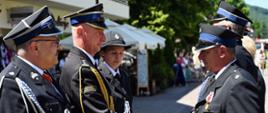 Na zdjęciu widać czterech mężczyzn i jedną kobietę w mundurach ochotniczej straży pożarnej, stojących podczas ceremonii. Dominują ciemne mundury z białymi koszulami i krawatami oraz czapkami z daszkiem. W centrum uwagi znajduje się dwóch mężczyzn po prawej stronie, którzy ściskają sobie dłonie, patrząc na siebie. Obaj mają na mundurach liczne odznaczenia i medale. Mężczyzna po lewej stronie tego uścisku, ma dodatkowo złoty sznur na ramieniu i pagonach. Jest to Śląski Komendant Wojewódzki PSP st. bryg. Damian Legierski. Za nimi, nieco po prawej, stoi młodsza kobieta również w mundurze. Trzyma w ręku płaską, srebrną tacę z medalami. W tle widać rozmyte otoczenie z zielenią drzew i krzewów, a także fragmenty budynków i parasoli, wydarzenie odbywa się na zewnątrz.