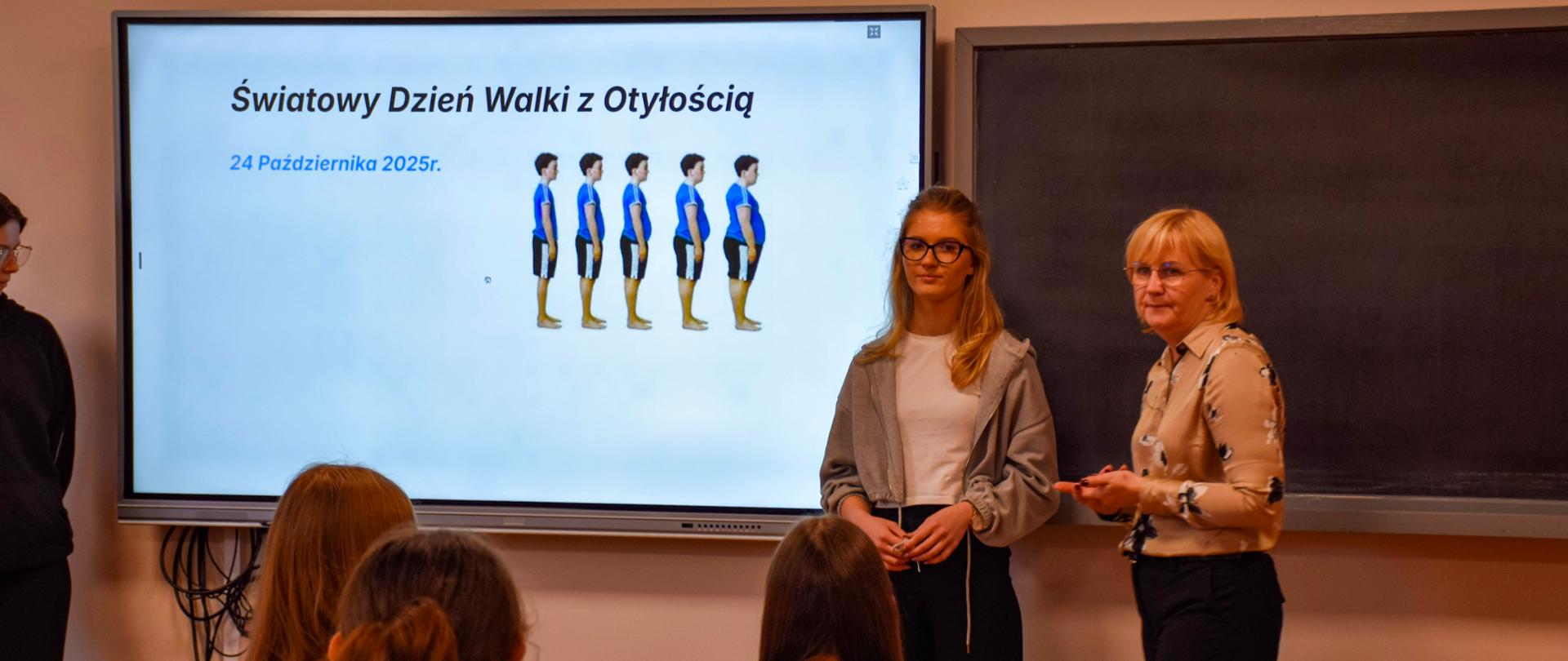 Plansza prezentacji o otyłości, obok prowadzące wykład kobiety - widok z tyłu klasy, zza pleców uczniów siedzących w ławkach