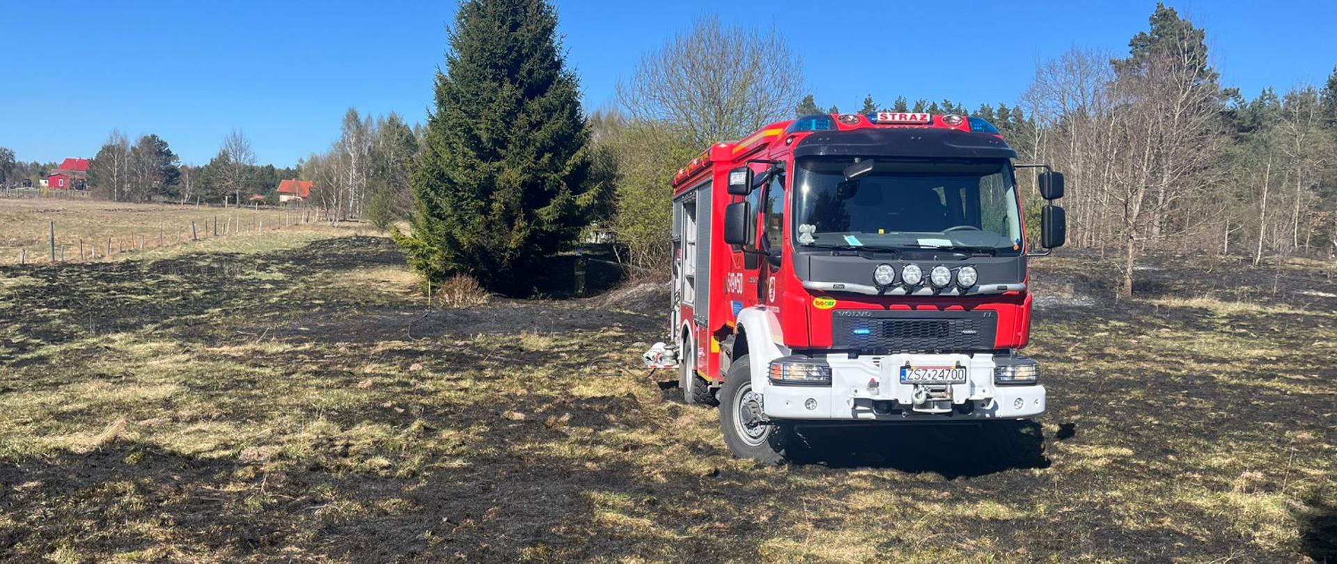 Na zdjęciu widać czerwony wóz strażacki stojący na pogorzelisku na otwartym terenie wiejskim. Trawa wokół jest częściowo spalona i pokryta czarnymi śladami po pożarze. W tle znajdują się drzewa oraz pojedyncze domy. Niebo jest bezchmurne i jasne, co sugeruje dobrą widoczność podczas akcji gaśniczej. Strażacy prawdopodobnie zakończyli lub kontrolują dogaszanie pożaru trawy.