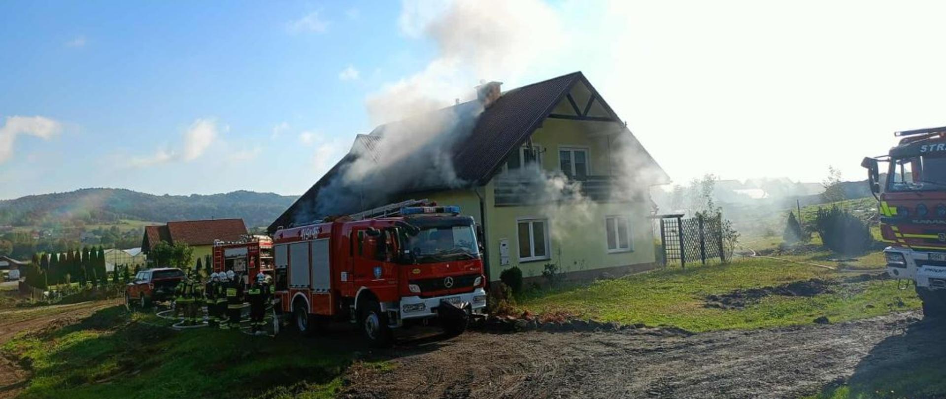 Kolorowa fotografia wykonana w pogodny dzień na zewnątrz. Przedstawia budynek mieszkalny z którego dachu i okien wydobywa się dym. Obok niego znajdują się samochody pożarnicze lub ich części przy których stoją strażacy w ubraniach specjalnych i hełmach z rozwiniętymi liniami wężowymi.