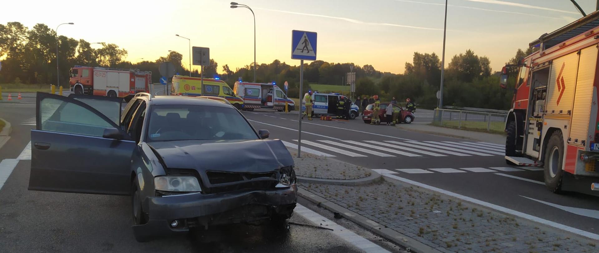 Zdjęcie przedstawia dwa pojazdy pożarnicze, pozostałe służby (radiowóz Policji i dwa Zespoły Ratownictwa Medycznego), pojazdy biorące udział w wypadku oraz strażaków prowadzących działania ratownicze