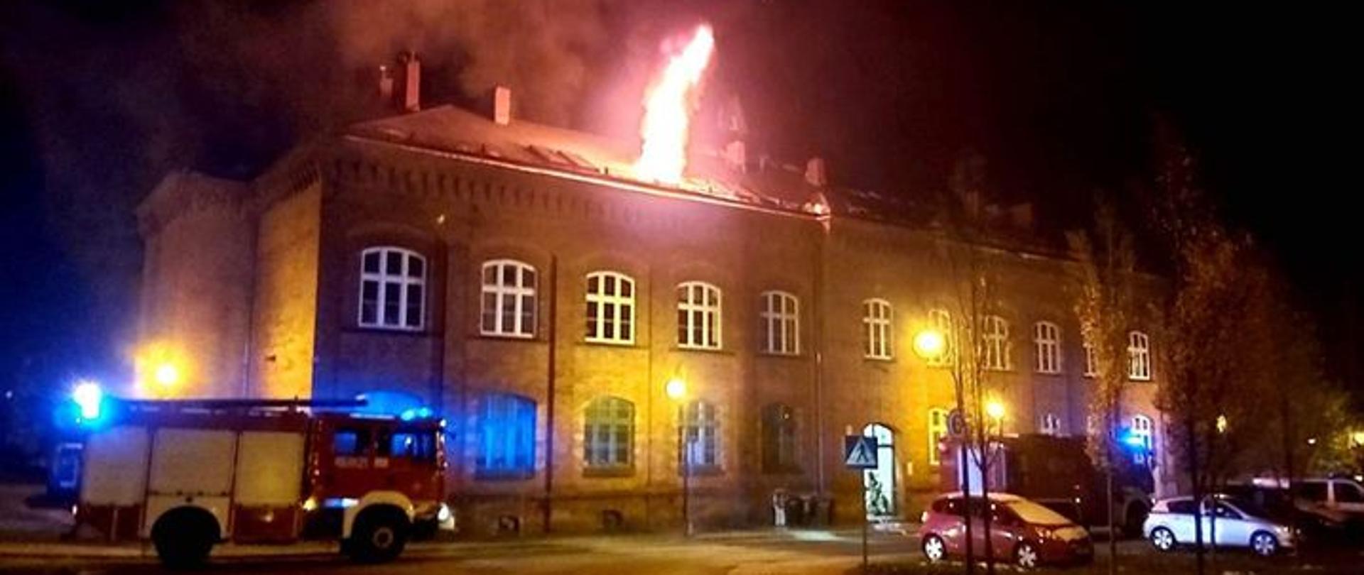 Płomień wydostaje się z dachu obiektu, przed którym stoi samochód straży pożarnej