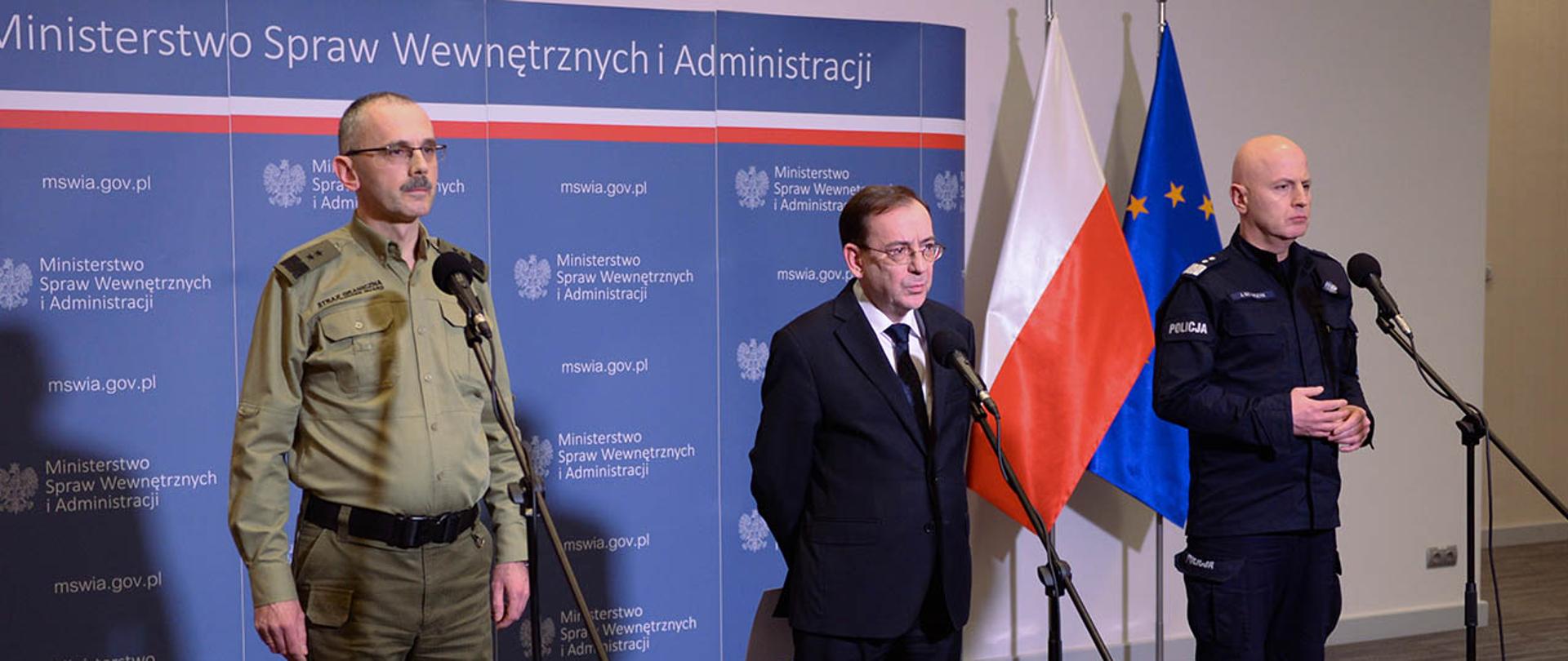 Na zdjęciu widać Mariusza Kamińskiego ministra SWiA, gen. dyw. SG Tomasza Pragę komendanta głównego Straży Granicznej i gen. insp. Jarosława Szymczyka komendanta głównego Policji stojących przy mikrofonach. W tle stoi ścianka MSWiA i flagi Polski i UE. 
