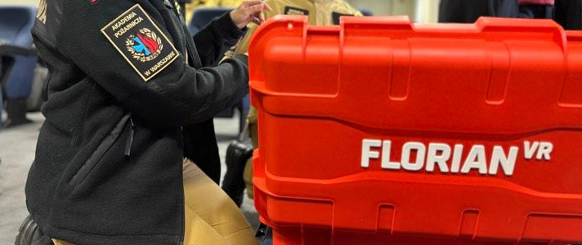 Strażacy podczas ćwiczeń z trenażerem Florian. Na pierwszym planie widoczna skrzynia transportowa oraz napis Florian. U strażaka po lewej stronie na ramieniu strażaka widoczny emblemat Akademii Pożarniczej w Warszawie.
