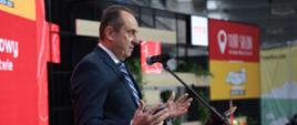 Wiceminister rozwoju i technologii Andrzej Gut-Mostowy przemawiający podczas otwarcia TOUR SALON w Poznaniu, wiceminister stoi za mównicą i trzyma uniesione dłonie