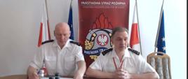 na zdjęciu widać obraz z kamery w trakcie wideokonferencji, zastępców łódzkiego Komendanta Wojewódzkiego PSP, w tle rollup Komendy Wojewódzkiej PPS w Łodzi oraz po lewej i prawej po 1 fladze Polski i unii europejskiej