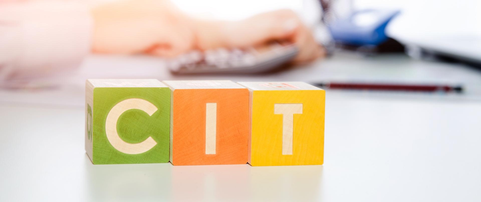 Napis na kolorowych klockach CIT a w tle rozmyty kalkulator i ludzkie dłonie.