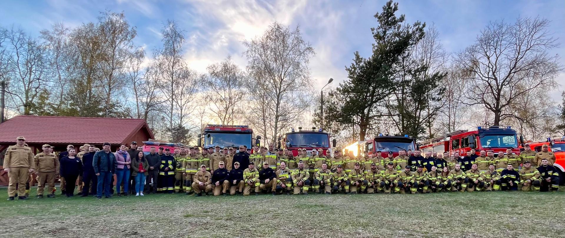 grupa strażaków w ubraniach specjalnych stojący w kilku rzędach, w tle pojazdy pożarnicze oraz drzewa 
