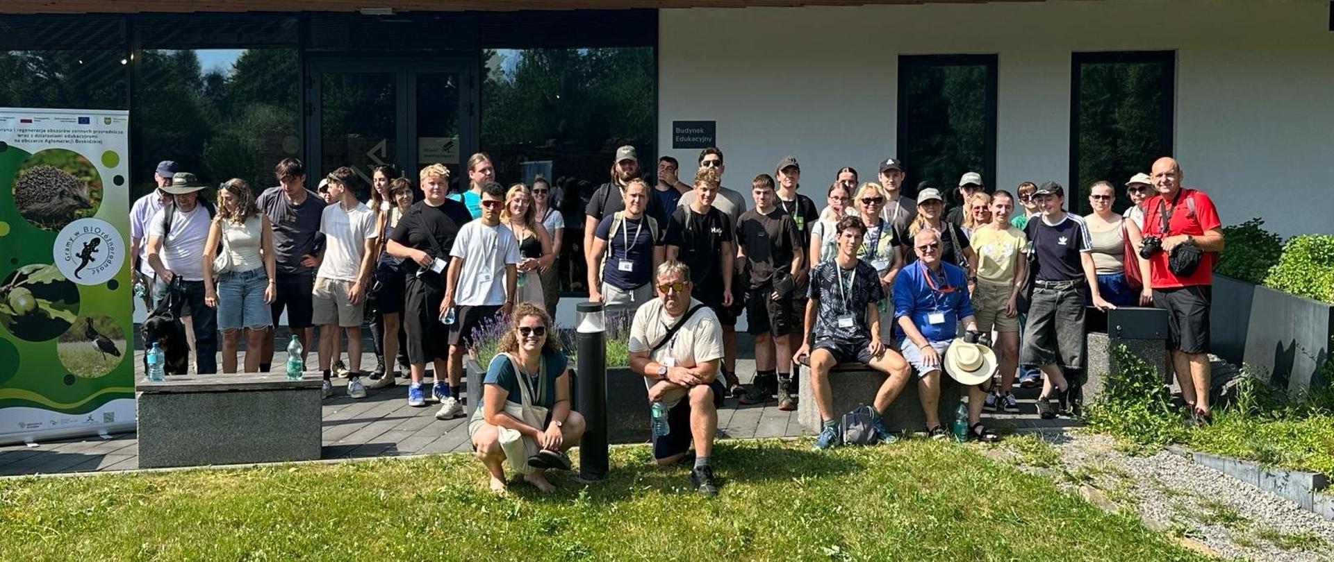 Grupa kilkudziesięciu osób stoi przed nowoczesnym budynkiem o metalowym dachu i dużych oknach. Uczestnicy pozują do wspólnego zdjęcia na trawniku; część ma identyfikatory na szyi. Po lewej stronie widoczny jest zielony baner z grafiką jaszczurki i tekstem związanym z biologią lub edukacją środowiskową. W tle rośliny ozdobne i wejście do budynku.
