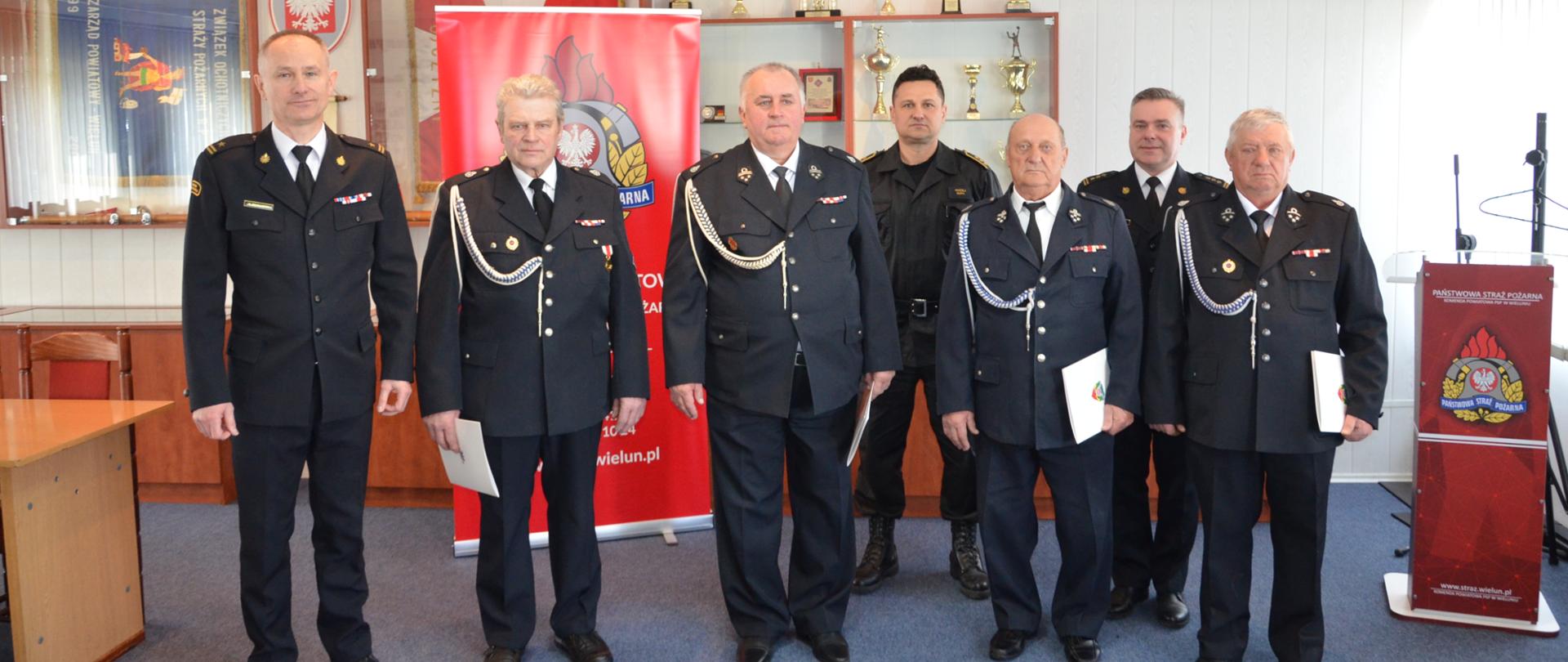 Zdjęcie przedstawia Komendanta Powiatowego Państwowej Straży Pożarnej w Wieluniu podczas wręczania decyzji o przyznaniu świadczenia ratowniczego strażakom Ochotniczych Straży Pożarnych w zbiorowym zdjęciu na tle ściany świetlicy konferencyjnej, widoczni także zastępca komendanta oraz funkcjonariusz do spraw kadr i czterech strażaków, którzy otrzymali świadczenie ratownicze, ubrani są w mundury wyjściowe