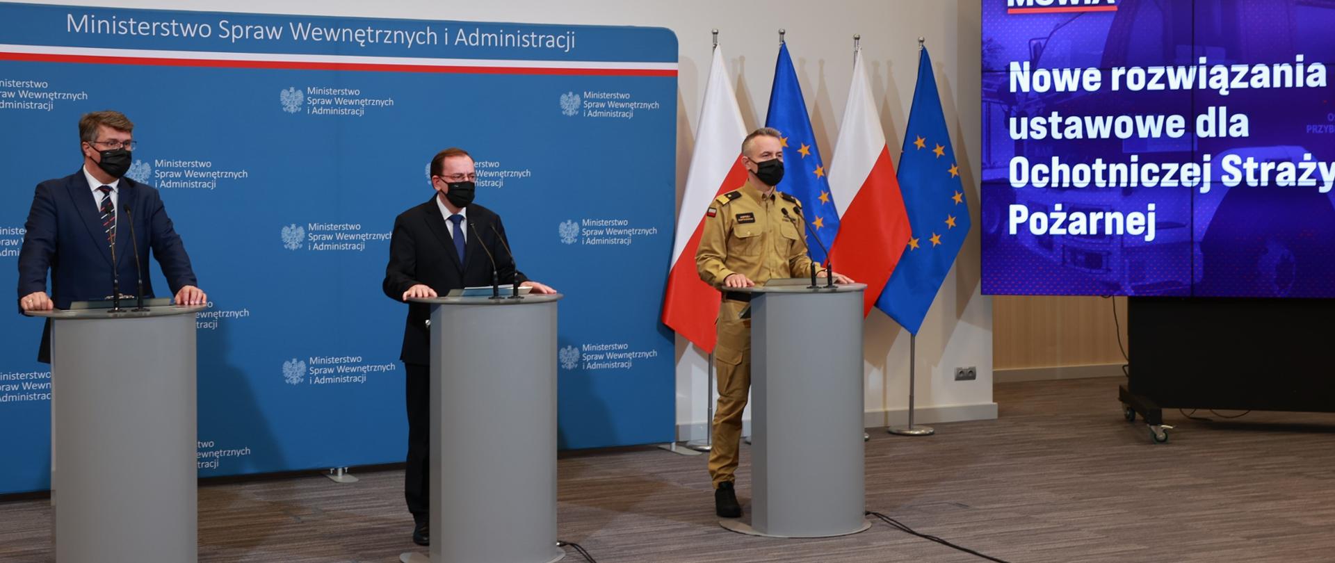 Na zdjęciu trzy osoby podczas konferencji prasowej stoją przy mównicach. W tle flagi Polski i UE oraz ścianka MSWiA