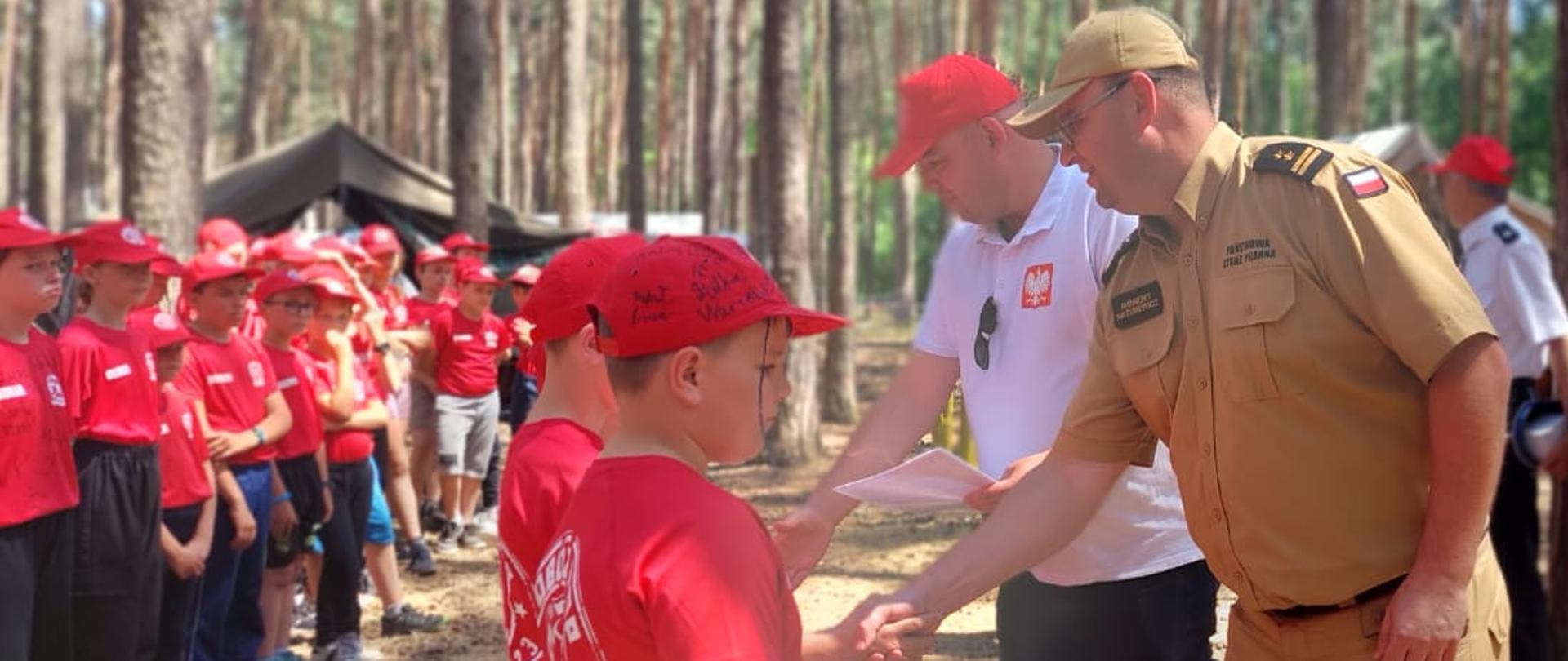 w lesie, strażak i mężczyzna w białej koszulce i czerwoinej czapeczce wręczają dyplomy druhom młodzieżowych drużym pożarniczych