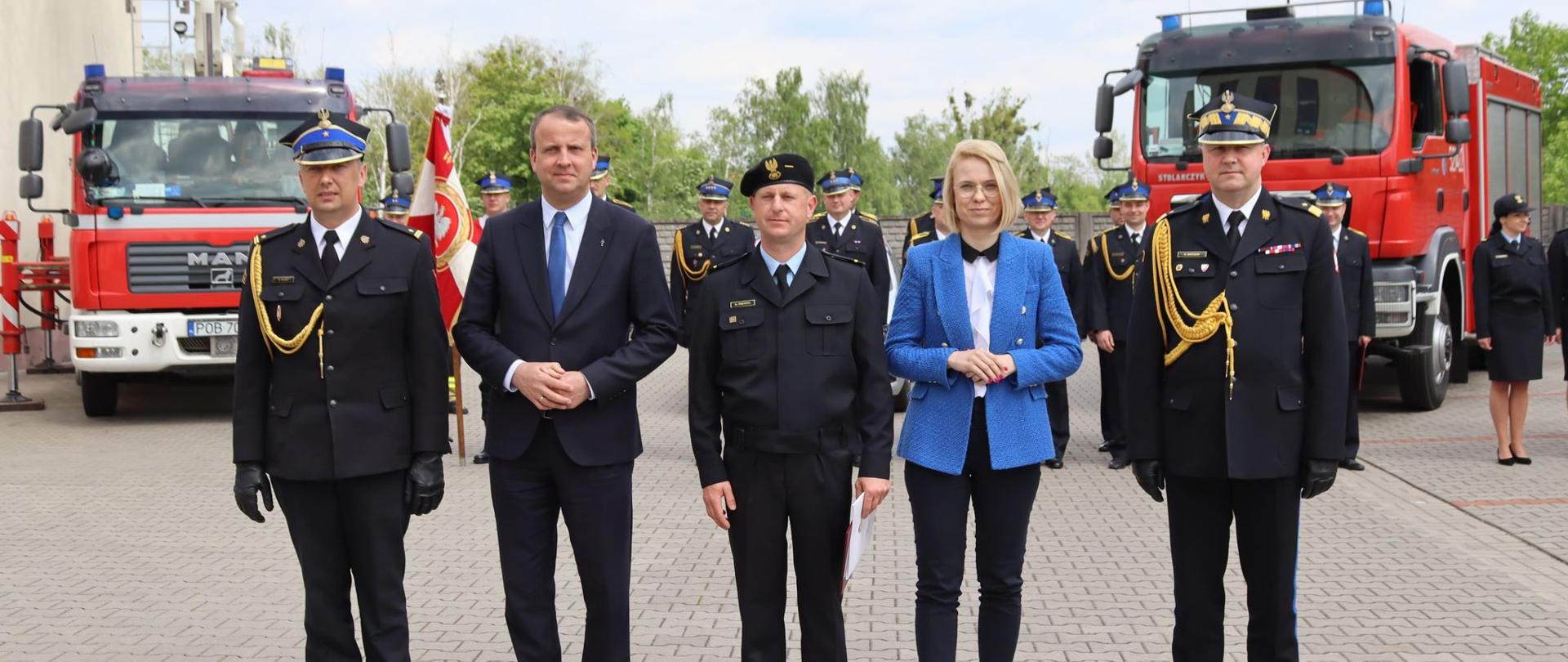 czterech mężczyzn w mundurach stoi w szeregu , w grupie stoi jedna kobieta, w tle stoją dwa czerwone samochody