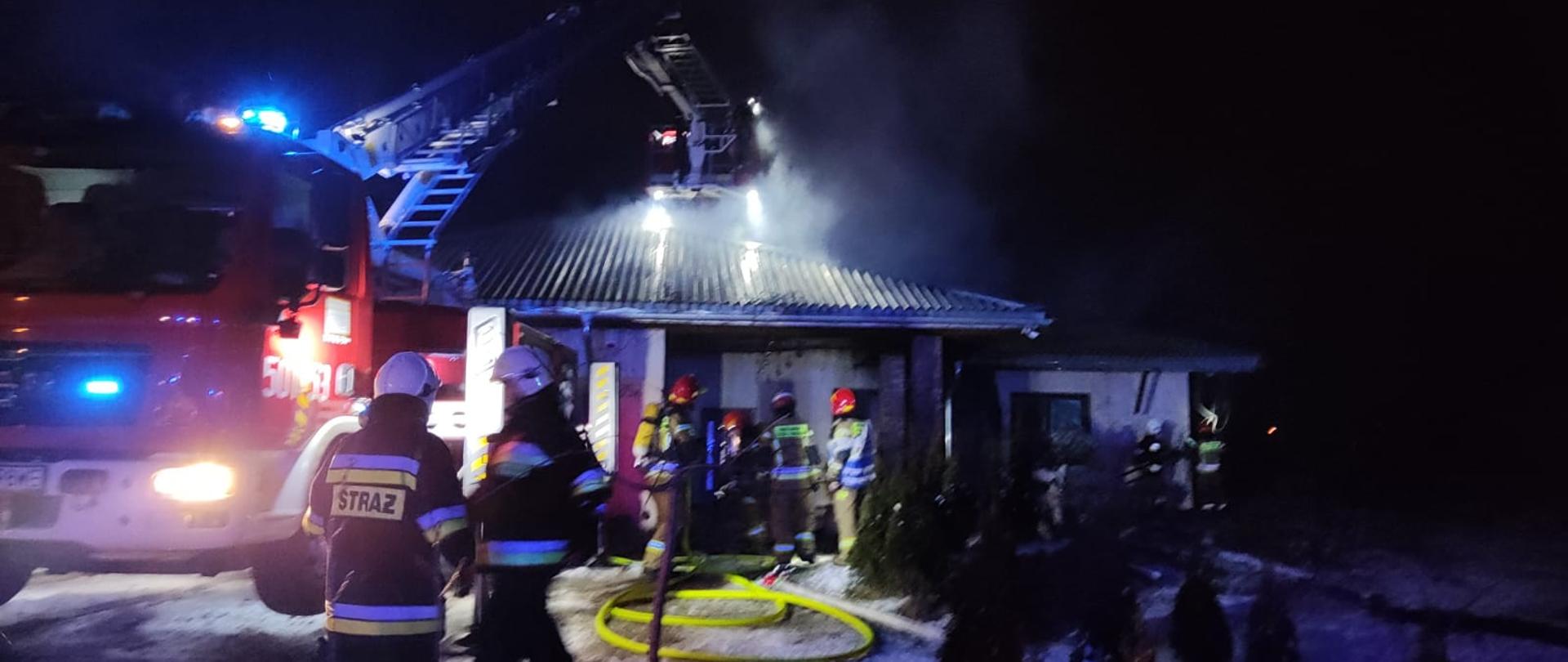 Czerwony strażacki wóz bojowy, widziany od przodu i częściowo z boku ma wyciągnięte do góry ramię z koszem, który jest oświetlony. Przy nim stoją dwaj strażacy ubrani w czarne mundury bojowe z białymi hełmami na głowie, obok nich rosną zielone tuje. W oddali stoli dom, z którego wydobywa się czarny dym. Przy wejściu do domu stoi czterech strażaków ubranych w piaskowe mundury bojowe i na głowach mają założone czerwone hełmy. Na plecach mają żółte butle z powietrzem, które służy do oddychania w zagrożonej strefie, wchodzą do budynku.