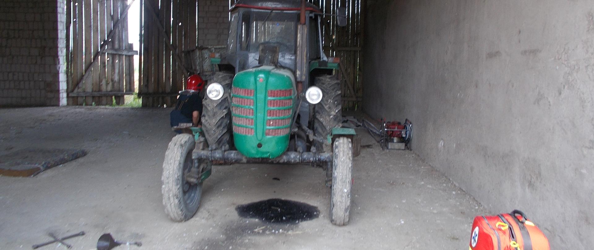 Garaż. Na betonowej posadzce stoi stary zielony ciągnik rolniczy z przyczepą. Pod ciągnikiem czarna plama oleju. Obok leżą klucz nasadowy oraz czarny lejek. Przy traktorze kuca strażak w ciemnym mundurze i czerwonym hełmie. W tle drewniane bramy garażu.
