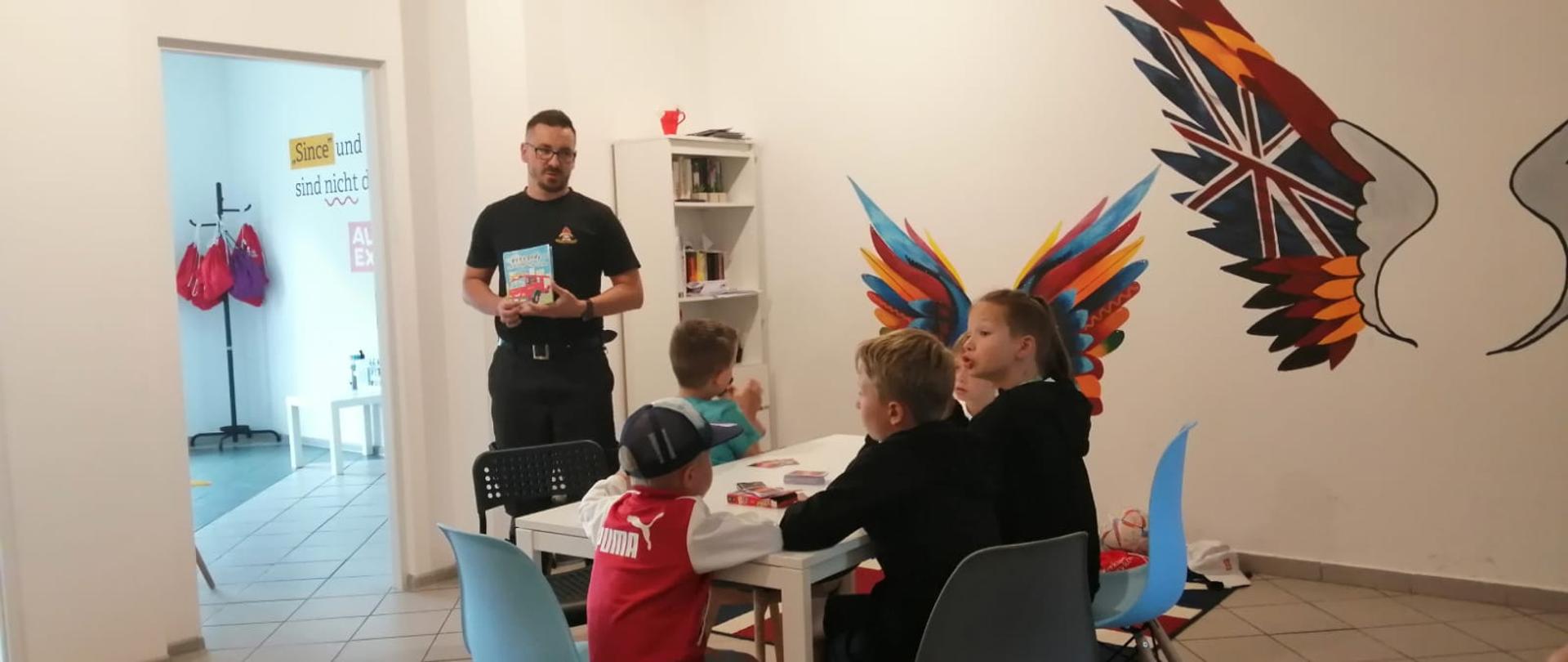 Strażak prowadzący pogadankę z dziećmi siedzącymi przy stole.