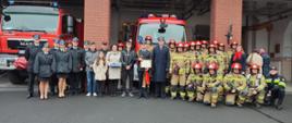 Strażacy w mundurach wyjściowych oraz specjalnych stoją przed garażem strażnicy, w tle samochody pożarnicze.