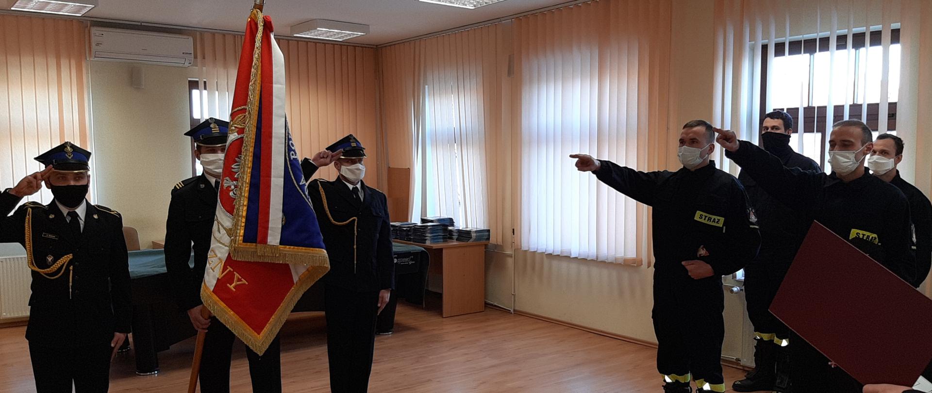 Poczet sztandarowy ze sztandarem, z boku 2 strażaków, którzy mówią rotę ślubowania. za nimi stoi kolejnych dwóch strażaków. 