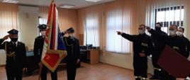 Poczet sztandarowy ze sztandarem, z boku 2 strażaków, którzy mówią rotę ślubowania. za nimi stoi kolejnych dwóch strażaków. 