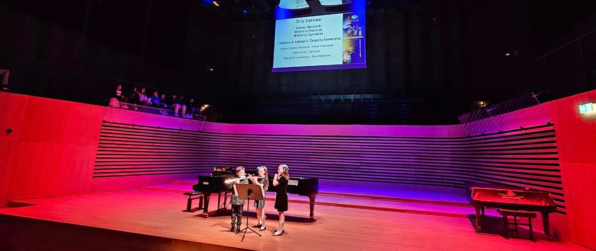 Chłopczyk i dwie dziewczynki grają na fletach na estradzie sali koncertowej, za nimi stoi fortepian.