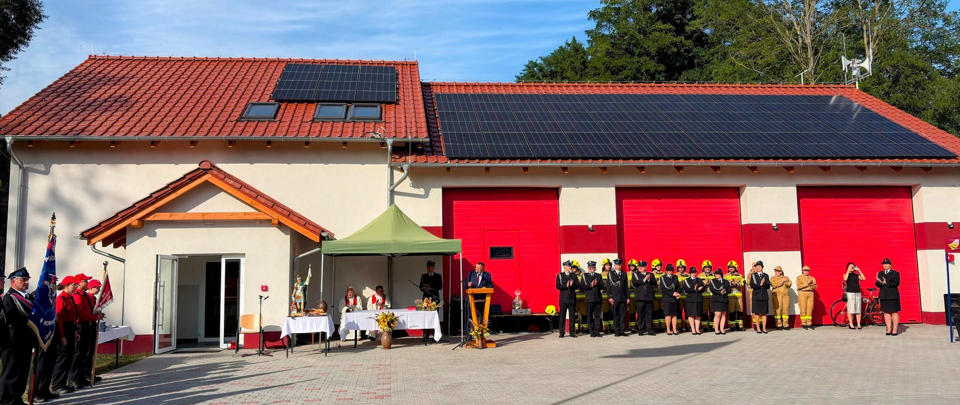 Nowa remiza OSP Łaz z trzema czerwonymi bramami garażowymi, na dachu zamontowane panele fotowoltaiczne. Przed budynkiem stoją pododdziały strażaków w mundurach galowych i bojowych, obok znajduje się scena z mównicą i stołem nakrytym białym obrusem.