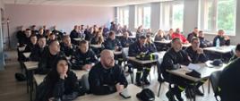 Strażacy (kobiety i mężczyźni) w czarnych mundurach siedzący przy dwuosobowych stołach w jasnej sali wykładowej