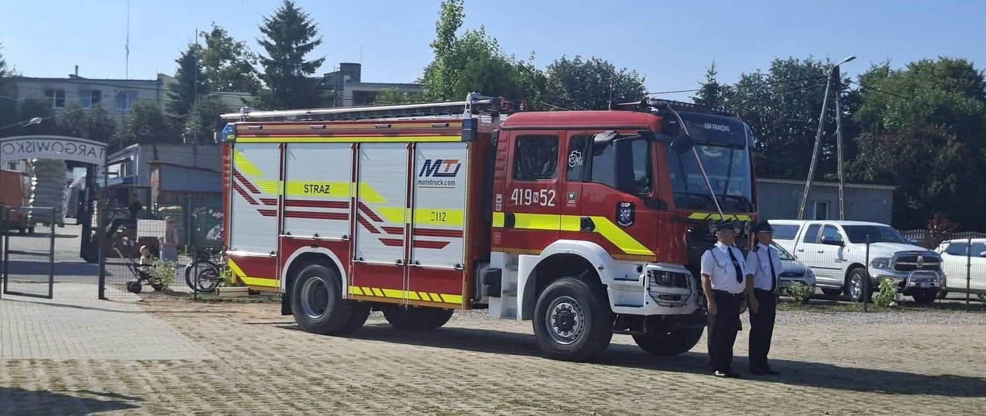 Na zdjęciu w centralnej części znajduje się pojazd ratowniczy gaśniczy, przed nim stoi dwóch druhów ubranych w mundur wyjściowy, czekają na oficjalne wręczenie kluczyków do pojazdu.