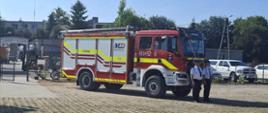Na zdjęciu w centralnej części znajduje się pojazd ratowniczy gaśniczy, przed nim stoi dwóch druhów ubranych w mundur wyjściowy, czekają na oficjalne wręczenie kluczyków do pojazdu.