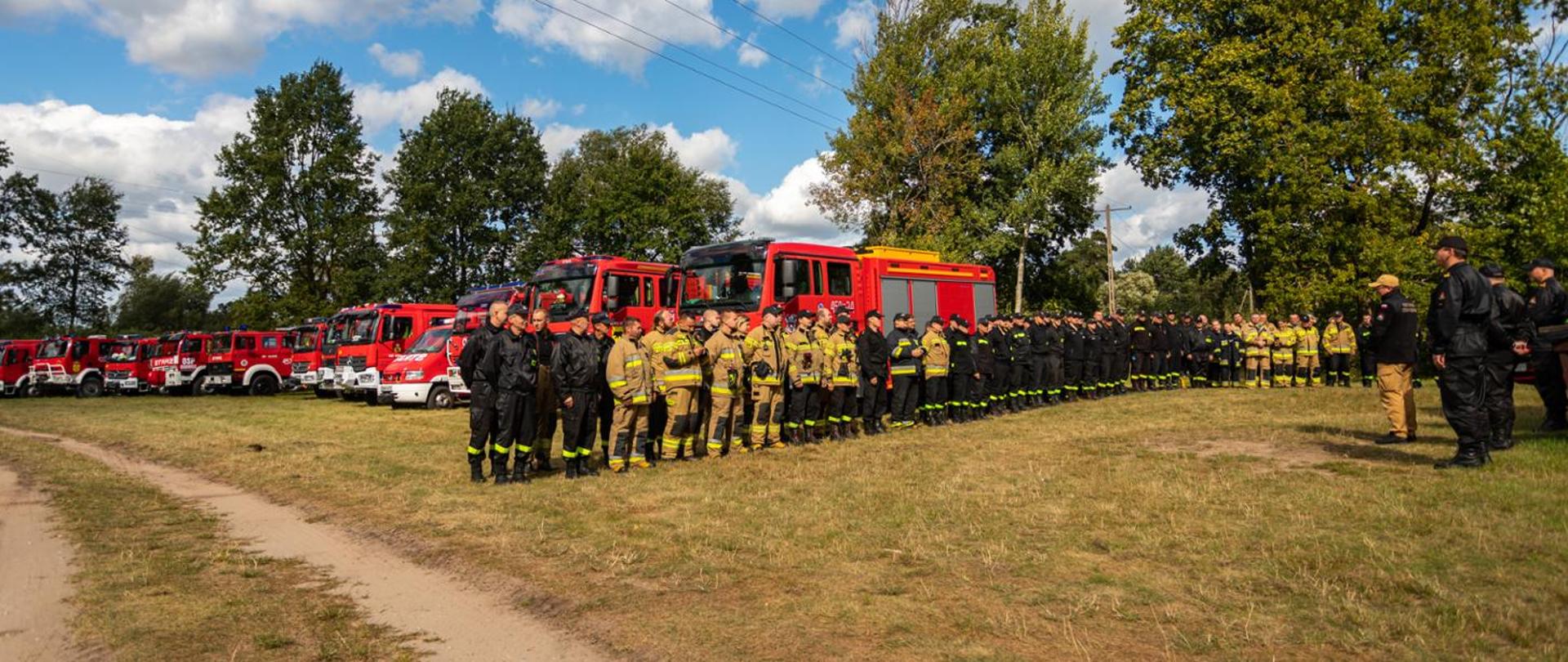 Na odprawie stoją w dwuszeregu strażacy PSP i OSP. W tle pojazdy strażackie.
